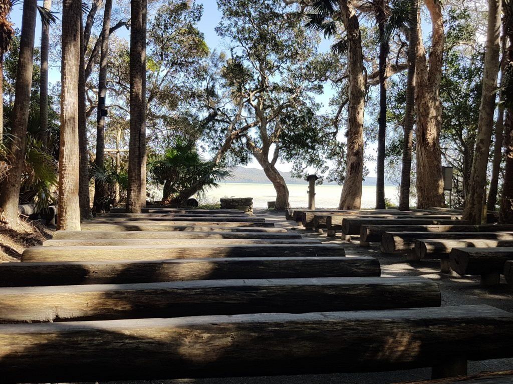 The Green Cathedral: rows of timber log pews under paperbarks and palms, open to Wallis Lake